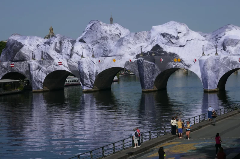 Bao · 5472 x 3648 Cette photographie montre un pont recouvert de sculptures géantes en forme de montagnes enneigées, créées avec un matériau blanc et brillant. Le sujet principal est cette installation artistique spectaculaire.La composition met en valeur le contraste entre les structures imposantes et l’eau calme en contrebas. Le cadrage inclut des personnes en bas, ajoutant une échelle humaine. La lumière est naturelle, avec un éclairage doux et légèrement bleuté, renforçant l’aspect hivernal. Les couleurs dominantes sont le blanc des sculptures et le gris-bleu de l’eau et du ciel.Le style est artistique et onirique, créant une ambiance à la fois fantastique et immersive.