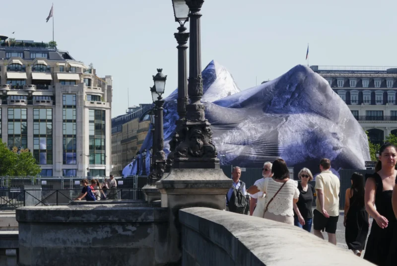 Bao · 5122 x 3439 Cette photographie met en scène une structure métallique recouverte d’un voile transparent bleu, évoquant une forme organique ou architecturale futuriste. Le sujet principal semble être cette installation artistique.La composition joue sur le contraste entre l’élément moderne et le pont historique en pierre. Le cadrage inclut des passants, ajoutant une dimension humaine et dynamique. La lumière est naturelle, avec un éclairage doux et homogène, suggérant une journée ensoleillée.Les couleurs dominantes sont les tons gris de la pierre et du métal, rehaussés par le bleu transparent de l’installation. L’ambiance est à la fois urbaine et artistique, mêlant tradition et innovation.