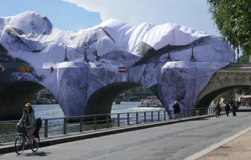 Bao · 5251 x 3361 Cette photographie montre une installation artistique monumentale représentant des blocs de glace géants, suspendus au-dessus d’un pont et d’un cours d’eau. Le sujet principal est cette œuvre éphémère, en tissu blanc et bleu pâle.La composition met en valeur l’architecture du pont et les passants, créant un contraste entre l’art et le quotidien. Le cadrage inclut une partie du ciel et des arbres, encadrant la scène. La lumière est naturelle, claire et ensoleillée, accentuant les teintes froides de l’installation. Les couleurs dominantes sont le blanc, le bleu et le gris, renforçant une ambiance à la fois poétique et mélancolique, évoquant la fragilité et la beauté éphémère.