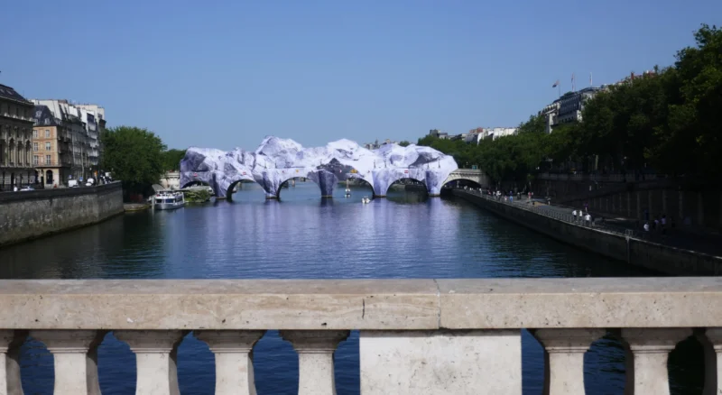 Bao · 5472 x 2993 Cette photographie montre un pont avec, en arrière-plan, une série d’arcs blancs suspendus au-dessus d’un canal, formant une structure artistique éphémère. Le sujet principal est cette installation lumineuse et architecturale.La composition est équilibrée, avec un cadrage horizontal large capturant le pont et le canal. La lumière est naturelle, claire et ensoleillée, mettant en valeur les structures. Les couleurs dominantes sont les bleus du ciel et de l’eau, contrastant avec les blancs des arcs. Le style est documentaire et esthétique, évoquant une ambiance à la fois moderne et festive.