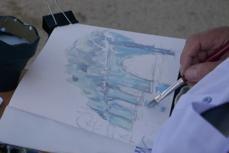 Bao · 5472 x 3648 Cette photographie montre une personne en train de dessiner ou peindre à l’aquarelle. Le sujet principal est une esquisse en cours, représentant une structure architecturale stylisée, probablement un pont ou un bâtiment.La composition est centrée sur le dessin, avec une main tenant un pinceau. Le cadrage est serré, mettant l’accent sur le processus créatif. La lumière semble naturelle et douce, éclairant uniformément la scène. Les couleurs dominantes sont les tons bleus et blancs, typiques de l’aquarelle. L’ambiance est calme et artistique, reflétant la concentration et la créativité.