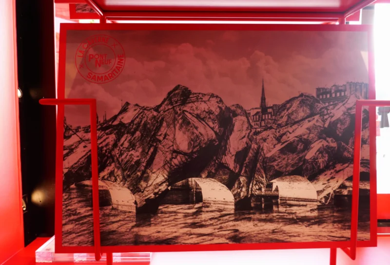 Bao · 5165 x 3512 Cette photographie montre une illustration encadrée représentant un paysage montagneux avec un village et un bateau sur une étendue d’eau.La composition est équilibrée, avec des montagnes en arrière-plan et un avant-plan aquatique. Le cadrage est centré, mettant en valeur les détails du dessin.Les couleurs dominantes sont des tons rouges et noirs, créant un style graphique et contrasté. La lumière semble artificielle, accentuant les contours et les ombres pour un effet dramatique.L’ambiance est à la fois mystérieuse et dramatique, renforcée par les teintes sombres et les jeux de lumière.