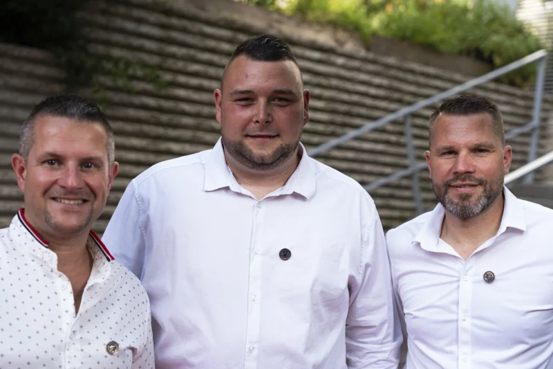 Cette photographie montre trois hommes posant côte à côte devant un escalier extérieur. Le sujet principal est leur présence souriante et détendue. La composition est symétrique, avec un cadrage serré sur les épaules et le haut du corps.La lumière semble naturelle, probablement en extérieur en journée, avec des ombres douces. Les couleurs dominantes sont les tons neutres : blanc des chemises, gris des vêtements et verts des plantes en arrière-plan. Le style est sobre et professionnel, évoquant une ambiance amicale et décontractée.