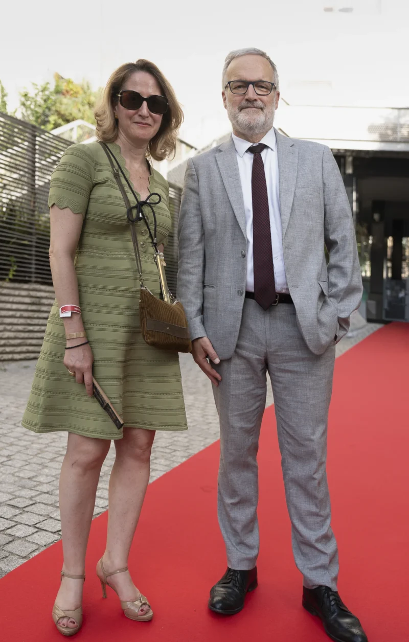 Cette photographie met en scène un couple sur un tapis rouge. La femme, vêtue d’une robe verte et de lunettes, tient un sac à main, tandis que l’homme porte un costume gris avec une cravate. Le cadrage est serré, centré sur eux deux, avec un arrière-plan flou et neutre.La lumière semble naturelle et douce, mettant en valeur leurs visages. Les couleurs dominantes sont les tons verts et gris, avec une touche de rouge due au tapis. L’ambiance est élégante et formelle, évoquant un événement mondain ou une cérémonie. Le style est sobre et sophistiqué.
