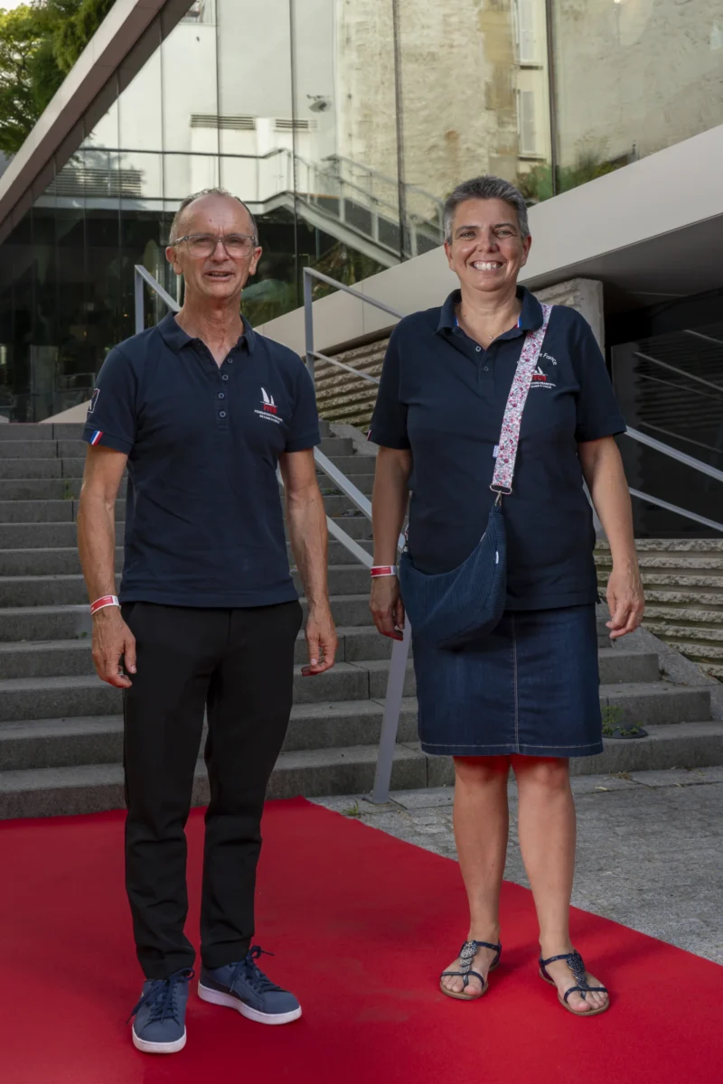 Cette photographie montre deux personnes debout sur un tapis rouge. Le sujet principal est un homme et une femme souriants, vêtus de tenues sportives bleues.La composition est symétrique, avec les deux sujets alignés verticalement. Le cadrage est serré, mettant en valeur les individus et les escaliers métalliques en arrière-plan. La lumière est naturelle et douce, éclairant uniformément les sujets.Les couleurs dominantes sont le bleu des vêtements, le rouge du tapis et les tons gris des escaliers. Le style est simple et naturel, évoquant une ambiance conviviale et professionnelle.