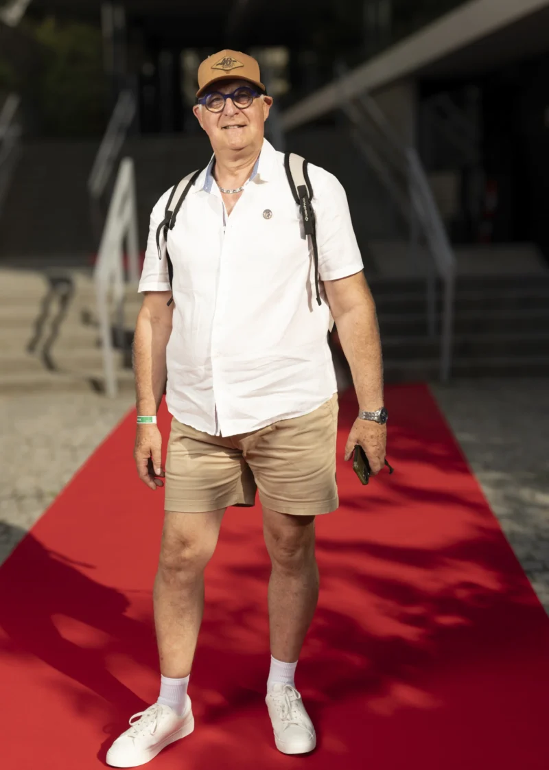 Cette photographie met en scène un homme comme sujet principal, vêtu d’une tenue décontractée (polo blanc, shorts, chapeau). Il se tient sur un tapis rouge, suggérant un événement formel.Le cadrage est serré, centré sur lui, avec un arrière-plan flou (structure métallique). La lumière est naturelle et douce, éclairant bien son visage. Les couleurs dominantes sont le blanc (vêtement), le beige (shorts), et le rouge vif du tapis, contrastant avec les tons neutres.Le style est simple et spontané, évoquant une ambiance à la fois élégante et décontractée, typique d’un événement public ou culturel.