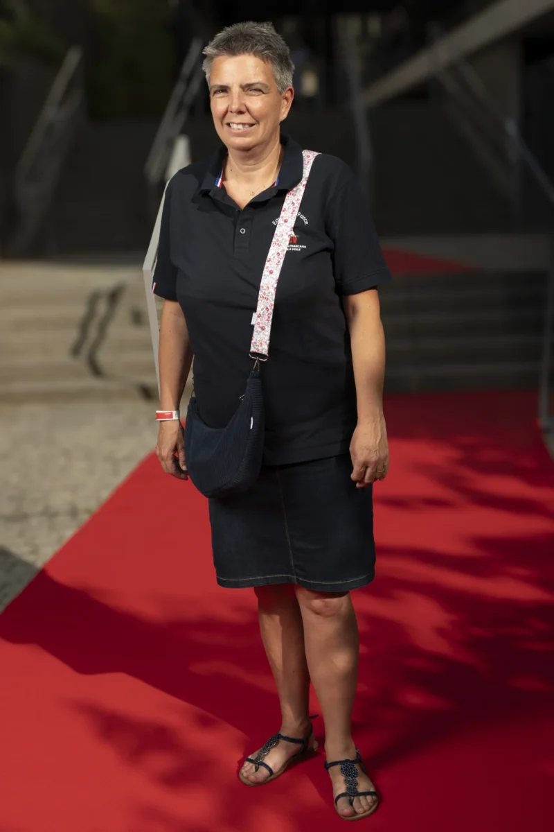 Cette photographie met en scène une femme comme sujet principal, vêtue d'une tenue sobre (robe noire et collier). Elle pose sur un tapis rouge, suggérant un événement formel.La composition est simple et centrée, avec un cadrage serré sur elle. L'arrière-plan flou, avec une structure métallique, attire peu l'attention. La lumière est naturelle et douce, éclairant son visage sans ombre marquée.Les couleurs dominantes sont le noir (sa tenue), le rouge (tapis) et des teintes métalliques froides. Le style est épuré et professionnel, créant une ambiance élégante et discrète.