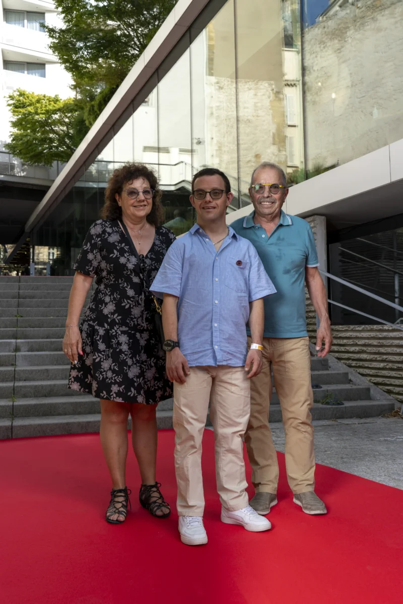 Cette photographie montre trois personnes posant sur un tapis rouge devant un bâtiment moderne. Le sujet principal est le trio, aligné et souriant.La composition est symétrique, avec un cadrage serré sur les individus. La lumière est naturelle et douce, éclairant uniformément les visages. Les couleurs dominantes sont les tons pastel de leurs vêtements (bleu, beige) et le rouge vif du tapis, contrastant avec le gris du bâtiment.Le style est sobre et professionnel, évoquant une ambiance formelle, probablement liée à un événement public ou une cérémonie.