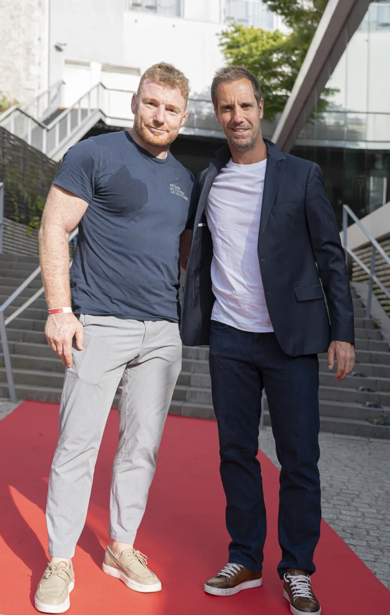 Cette photographie montre deux hommes posant côte à côte sur un tapis rouge. Le sujet principal est leur présence amicale, habillés de manière décontractée mais élégante.La composition est symétrique, avec un cadrage serré sur les deux individus. L’arrière-plan, flou, révèle une structure métallique et des escaliers, suggérant un lieu urbain moderne. La lumière est naturelle et douce, éclairant uniformément les sujets. Les couleurs dominantes sont les tons bleus de leurs vêtements, contrastant avec le rouge vif du tapis et les gris métalliques de l’environnement. L’ambiance est à la fois professionnelle et détendue.