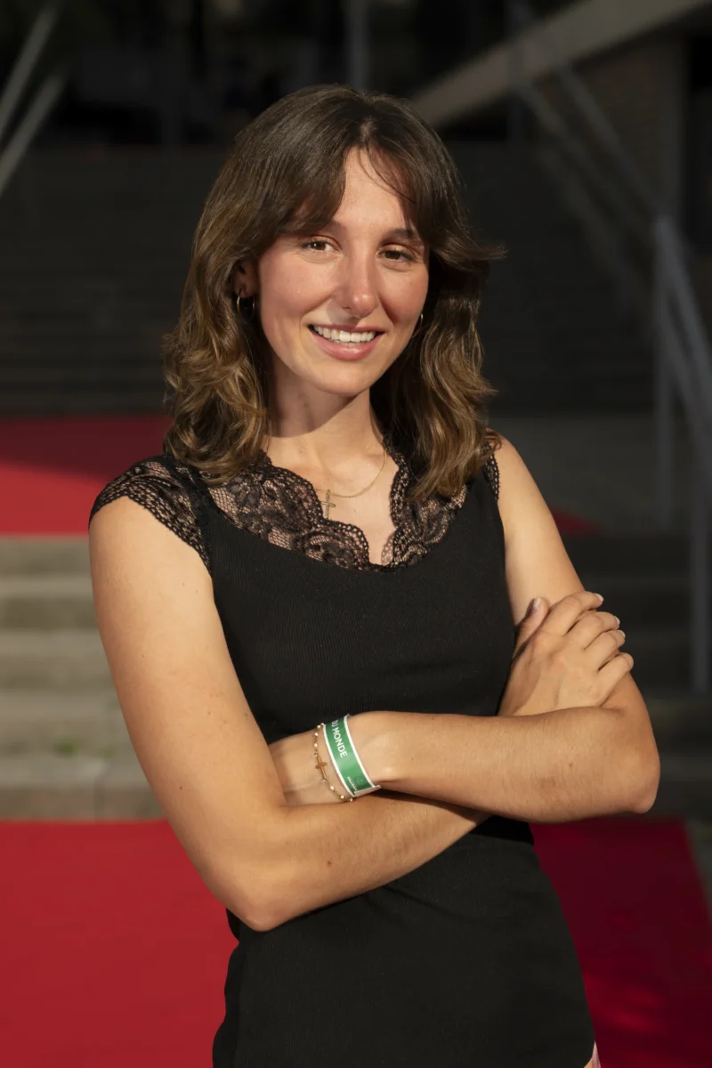 Cette photographie met en scène une femme comme sujet principal, souriante et posant avec les bras croisés. La composition est simple et centrée, avec un cadrage serré sur elle.La lumière est douce et naturelle, éclairant son visage sans ombres marquées. Les couleurs dominantes sont les tons chauds : noir de sa tenue, rouge du fond, et touches de vert sur ses bracelets.Le style est sobre et élégant, évoquant une ambiance professionnelle ou événementielle. L'atmosphère est chaleureuse et accueillante.