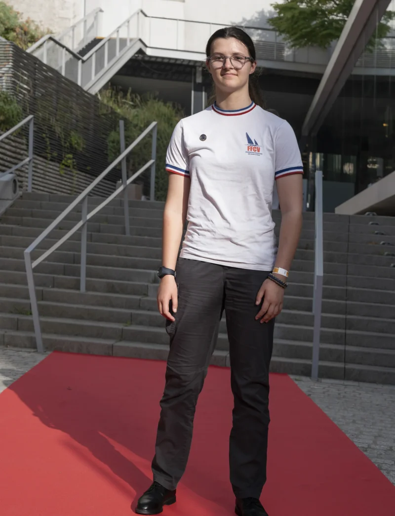 Cette photographie met en scène un jeune homme comme sujet principal, debout sur un tapis rouge. Il porte un t-shirt blanc à manches courtes et un pantalon gris.La composition est simple, avec un cadrage serré sur le sujet, les escaliers métalliques en arrière-plan. La lumière semble naturelle, douce et légèrement latérale, créant des ombres subtiles. Les couleurs dominantes sont les tons neutres (gris, blanc) et le rouge vif du tapis.Le style est sobre et contemporain, évoquant une ambiance formelle mais détendue, peut-être liée à un événement public ou une cérémonie.