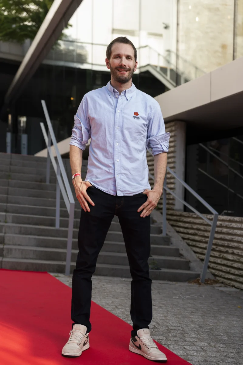 Cette photographie met en scène un homme comme sujet principal, debout sur un tapis rouge. Il porte une chemise légère et un pantalon sombre, avec des chaussures décontractées.La composition est simple et centrée, avec un cadrage serré sur le sujet. L’arrière-plan architectural, avec des escaliers et des structures en béton, suggère un lieu public ou un événement formel.La lumière est naturelle et douce, éclairant bien le sujet sans ombres marquées. Les couleurs dominantes sont les tons neutres (gris, bleu clair) et le rouge vif du tapis, créant un contraste élégant. L’ambiance est à la fois décontractée et légèrement formelle.