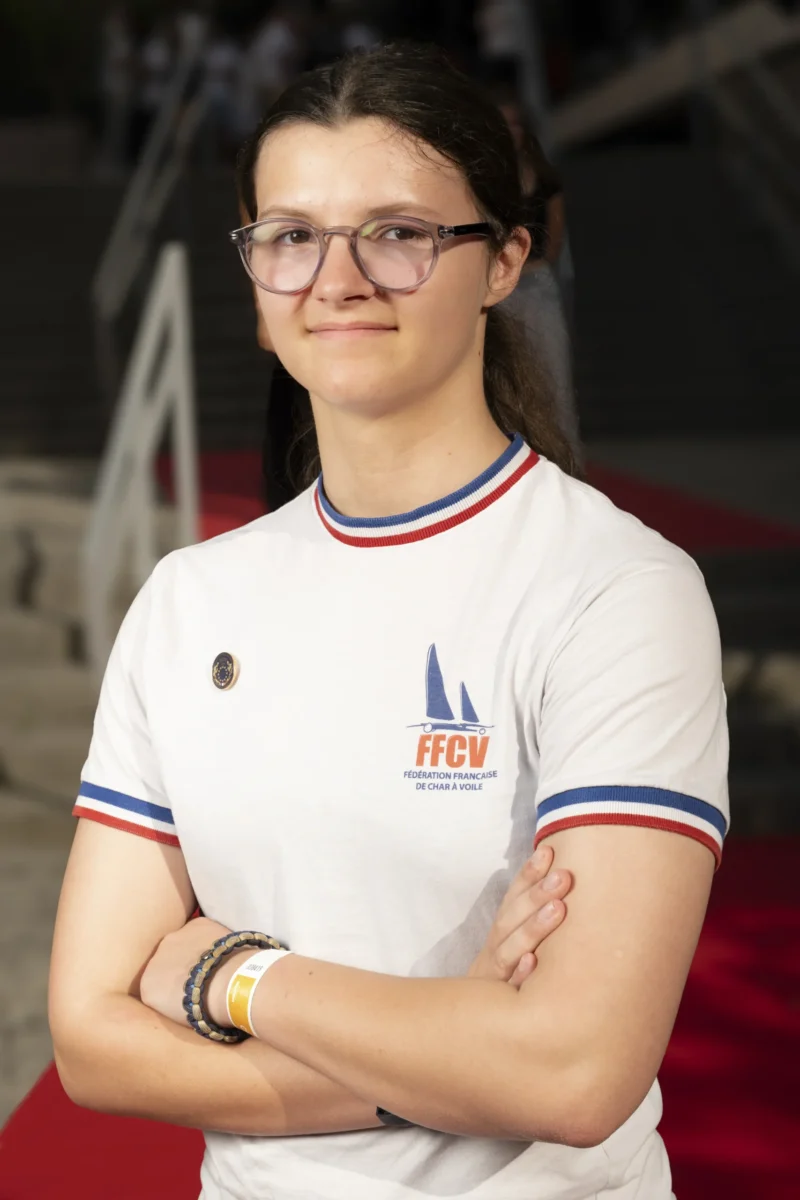 Cette photographie met en scène une jeune femme comme sujet principal, vêtue d’un t-shirt blanc aux couleurs et logo de la FFGV (Fédération Française de Gymnastique Volontaire). Elle pose les bras croisés, avec un cadrage serré centré sur elle. La lumière est douce et uniforme, éclairant son visage et ses épaules sans ombre marquée. Les couleurs dominantes sont le blanc du t-shirt, rehaussé de bleu, rouge et noir pour les détails. Le style est sobre et sportif, évoquant une ambiance professionnelle et déterminée.