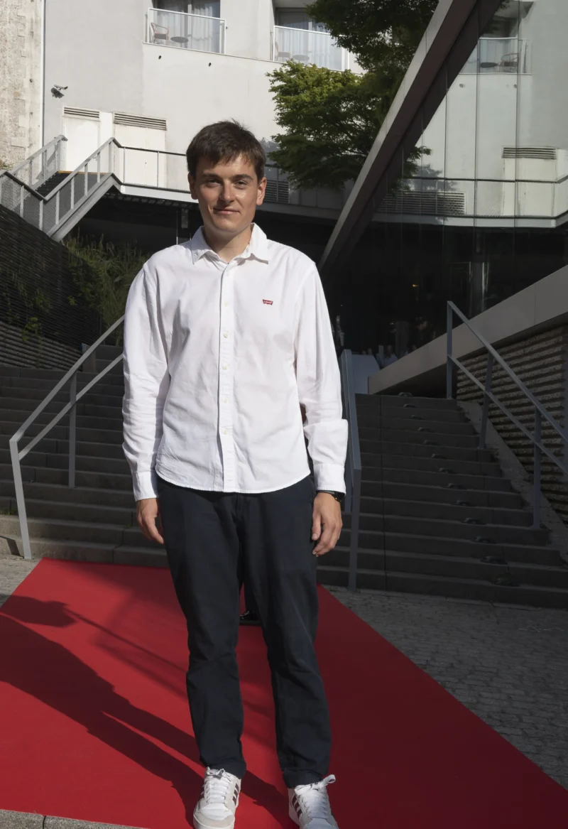 Cette photographie montre un homme en tenue décontractée (veste blanche, pantalon noir et baskets) comme sujet principal. Il se tient sur un tapis rouge, devant un escalier extérieur menant à un bâtiment moderne.Le cadrage est serré, mettant l’accent sur lui. La lumière est naturelle, probablement en journée, avec des ombres douces. Les couleurs dominantes sont le blanc (vêtement), le rouge (tapis), le gris (bâtiment) et le vert (végétation en arrière-plan). Le style est sobre et contemporain, évoquant une ambiance professionnelle ou événementielle, malgré la simplicité du sujet.