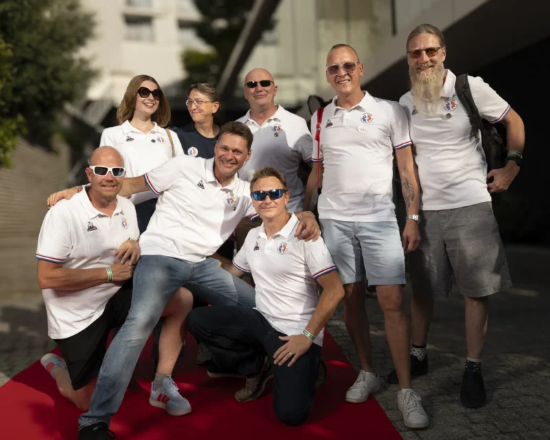 Cette photographie montre un groupe de sept personnes posant sur un tapis rouge. Le sujet principal est l'ensemble du groupe, vêtu de tenues blanches assorties, avec des lunettes de soleil.La composition est symétrique, avec les individus disposés en deux rangées. Le cadrage est serré, mettant en valeur les visages et les expressions. La lumière est naturelle et douce, créant des ombres légères. Les couleurs dominantes sont le blanc des vêtements, le rouge du tapis et les tons neutres des visages et du fond.L’ambiance est décontractée et festive, évoquant un événement informel ou une célébration.