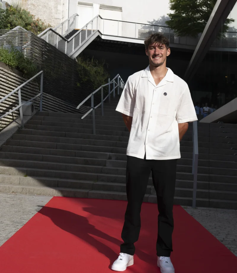 Cette photographie met en scène un jeune homme comme sujet principal, souriant et debout sur un tapis rouge. La composition est simple et centrée, avec des escaliers métalliques en arrière-plan.Le cadrage est serré, mettant l’accent sur le sujet. La lumière est naturelle et douce, éclairant bien son visage. Les couleurs dominantes sont le blanc (son vêtement), le rouge (le tapis) et les tons neutres des escaliers. Le style est épuré et contemporain, évoquant une ambiance formelle mais décontractée, peut-être liée à un événement public ou une cérémonie.