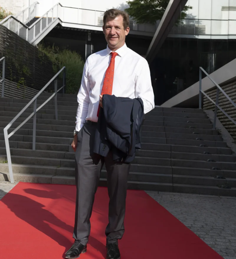 Cette photographie montre un homme en costume-cravate comme sujet principal, souriant et posant sur un tapis rouge. La composition met en valeur les escaliers et une rampe en arrière-plan. Le cadrage est serré sur le sujet, avec une légère inclinaison vers le haut.La lumière semble naturelle, bien répartie, sans ombres marquées. Les couleurs dominantes sont les tons neutres du costume gris, le rouge vif du tapis et le blanc de la chemise. L’ambiance est formelle et élégante, évoquant un événement officiel ou une cérémonie.
