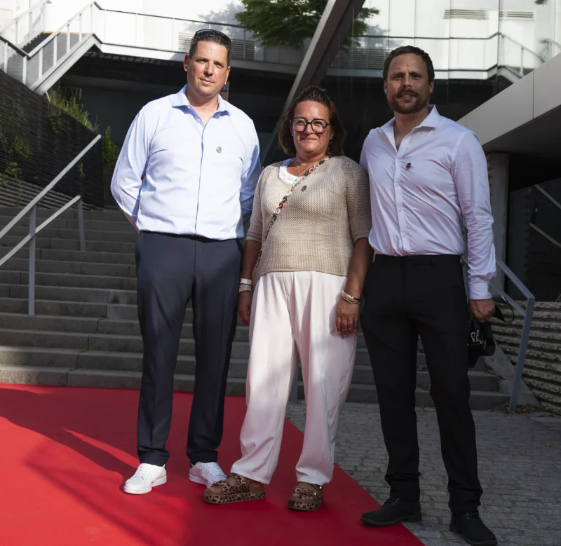 Cette photographie montre trois personnes posant sur un tapis rouge. Le sujet principal est ce trio, composé de deux hommes et une femme, habillés élégamment.La composition est symétrique, avec les individus alignés verticalement. Le cadrage est serré, mettant l'accent sur eux. La lumière semble naturelle, douce et uniforme, sans ombres marquées. Les couleurs dominantes sont les tons neutres (gris, beige) et les touches de bleu et blanc dans les vêtements. L’ambiance est formelle et sophistiquée, évoquant un événement public ou une cérémonie.