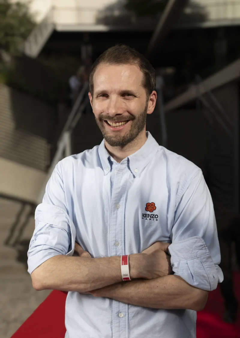 Cette photographie met en scène un homme souriant, sujet principal, vêtu d'une chemise légère à manches roulées avec un logo visible. Il pose les bras croisés sur un tapis rouge, devant un fond flou avec une structure métallique.Le cadrage est serré, centré sur lui, avec une lumière naturelle douce et homogène. Les couleurs dominantes sont les tons clairs de sa chemise (bleu pâle) et le rouge du tapis, créant un contraste chaleureux. L’ambiance est professionnelle et décontractée, évoquant un événement ou une interview. Le style est sobre et moderne.