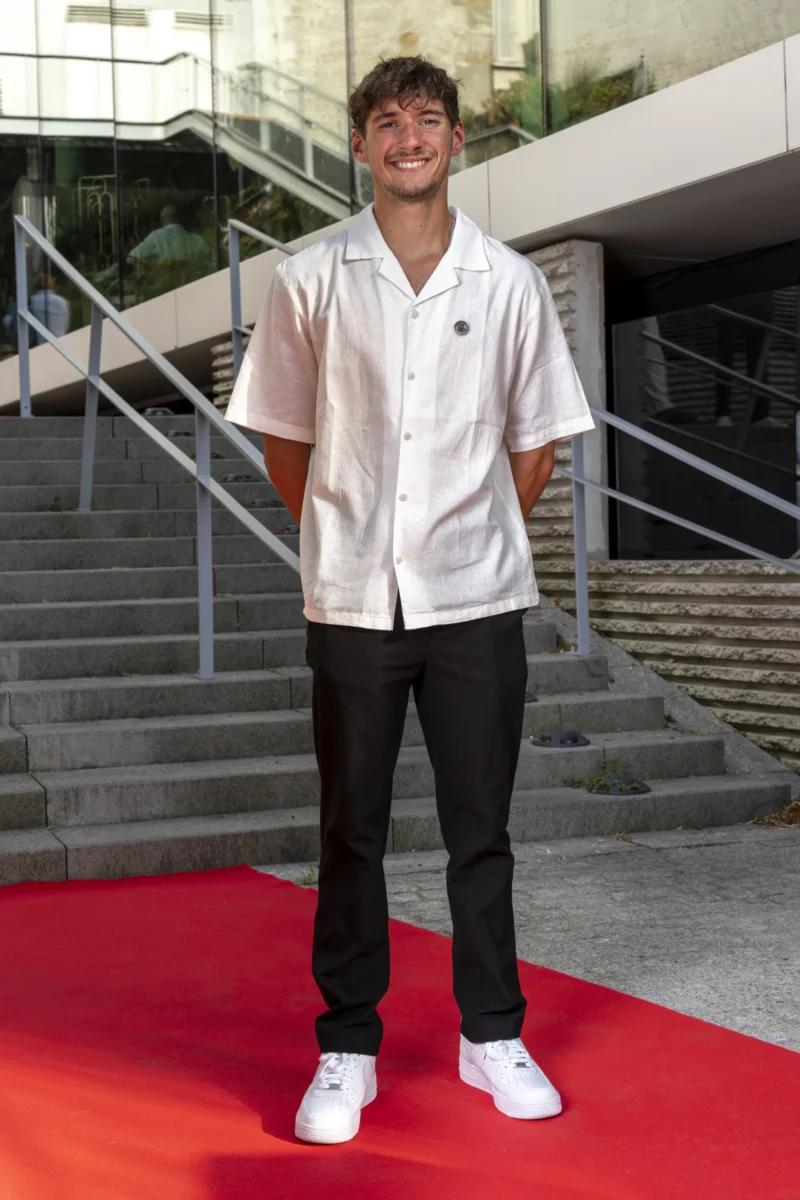 Cette photographie met en scène un jeune homme en tenue blanche (chemise et pantalon) avec des baskets blanches, debout sur un tapis rouge. Le sujet principal est bien cadré, centré et légèrement décalé vers la gauche. L’arrière-plan montre des escaliers et une structure architecturale moderne.La lumière est naturelle et douce, éclairant le sujet de face. Les couleurs dominantes sont le blanc (vêtements), le rouge (tapis) et les tons neutres de l’architecture. Le style est épuré et professionnel, évoquant une ambiance formelle, peut-être liée à un événement ou une présentation.