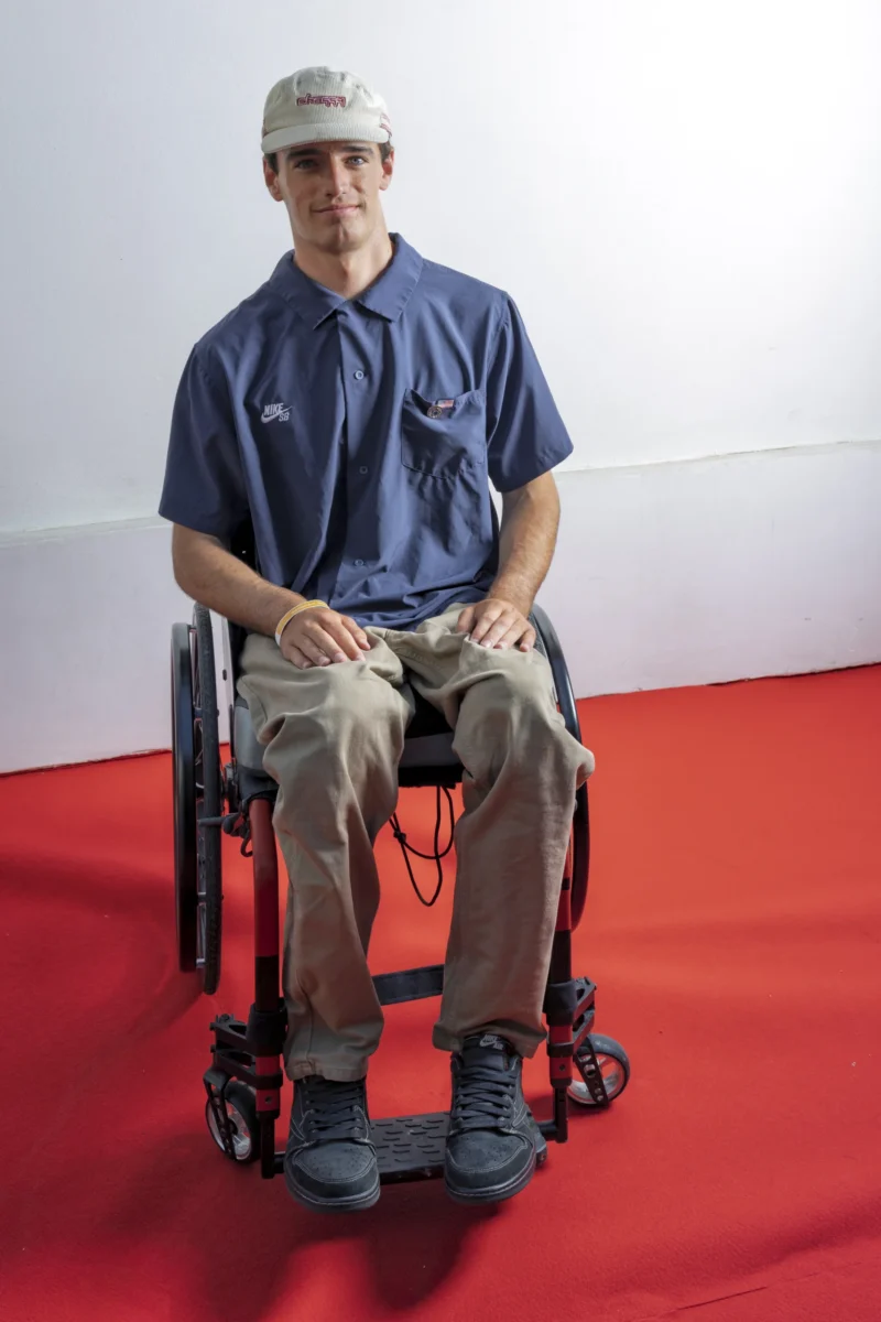 Cette photographie montre un homme assis dans un fauteuil roulant comme sujet principal. La composition est simple et centrée, avec un cadrage serré mettant en valeur le sujet. La lumière est douce et uniforme, éclairant clairement son visage et ses vêtements.Les couleurs dominantes sont les tons bleus de son polo, les beige et gris de son pantalon, ainsi que le rouge vif du tapis au sol. Le style est réaliste et documentaire. L’ambiance est calme et sobre, mettant en avant une touche d’humanité et de simplicité.