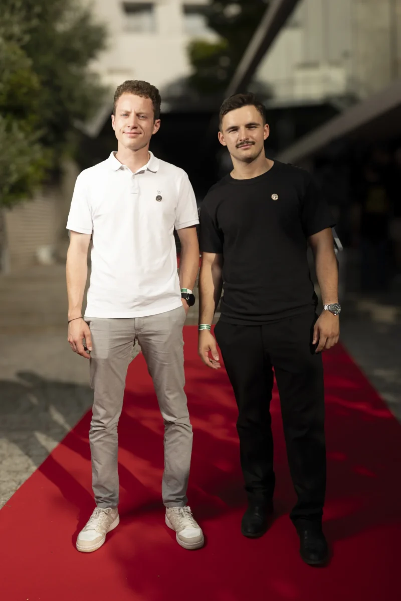 Cette photographie montre deux hommes debout sur un tapis rouge. Le sujet principal est ces deux individus, l’un vêtu d’un polo blanc et l’autre d’un t-shirt noir.La composition est symétrique, avec un cadrage serré sur les deux hommes. La lumière est naturelle et douce, éclairant leurs visages sans ombres marquées. Les couleurs dominantes sont les tons neutres (noir, blanc, gris) et le rouge vif du tapis.Le style est sobre et professionnel, évoquant une ambiance formelle, probablement liée à un événement public ou une cérémonie.