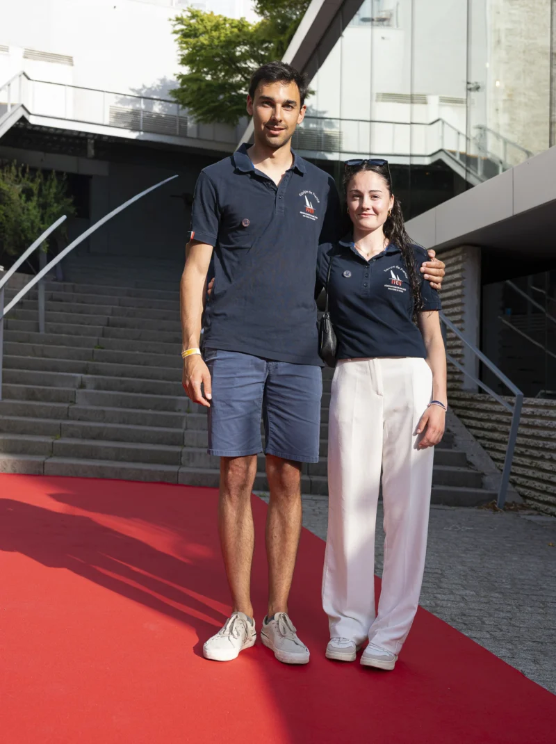Cette photographie met en scène deux personnes posant côte à côte sur un tapis rouge. Le sujet principal est un homme et une femme souriants, vêtus de tenues sportives (un polo et un short pour lui, un survêtement pour elle).La composition est symétrique, avec un cadrage serré sur les deux individus. L’arrière-plan montre des escaliers et une structure architecturale moderne. La lumière est naturelle et douce, éclairant uniformément les sujets. Les couleurs dominantes sont les tons neutres (gris, blanc) et les couleurs vives du tapis rouge. L’ambiance est décontractée et joyeuse, évoquant un événement sportif ou informel.