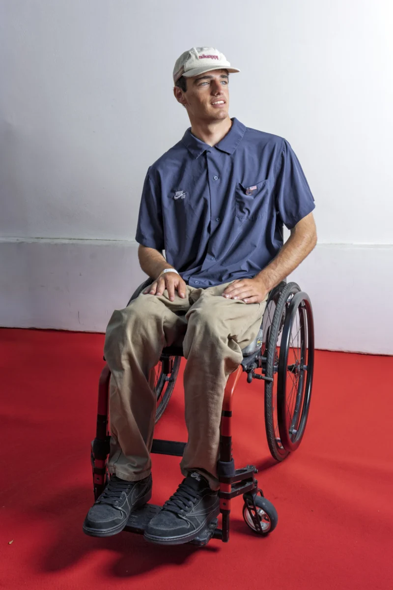 Cette photographie met en scène un homme assis dans un fauteuil roulant, sujet principal central. Le cadrage est serré, le plaçant devant un fond uni blanc et rouge.La lumière est douce et uniforme, éclairant clairement son visage et ses vêtements. Les couleurs dominantes sont le bleu (vêtement), le beige (pantalon), le rouge (sol) et le blanc (arrière-plan). Le style est sobre et direct, avec une ambiance calme et neutre, mettant l'accent sur la personne sans distraction.