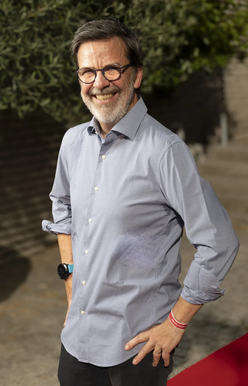 Cette photographie met en scène un homme souriant comme sujet principal, vêtu d'une chemise bleu clair à manches retroussées. Le cadrage est serré, centré sur lui, avec un fond légèrement flou de végétation.La lumière est naturelle et douce, éclairant son visage de manière homogène. Les couleurs dominantes sont les tons bleus de sa chemise, contrastant avec les verts du fond. L’ambiance est chaleureuse et détendue, évoquant une scène informelle et positive. Le style est simple et direct, sans artifices.