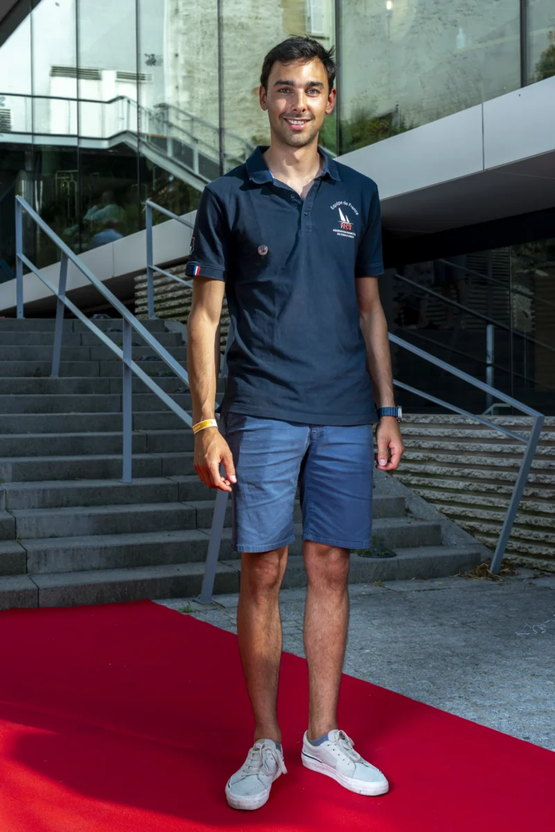 Cette photographie met en scène un jeune homme en tenue sportive, polo bleu et shorts assortis, posant sur un tapis rouge. Le sujet principal est bien cadré, centré et légèrement de trois quarts.L’arrière-plan montre des escaliers et une structure architecturale moderne. La lumière est naturelle et douce, éclairant le sujet de face. Les couleurs dominantes sont le bleu du vêtement, le rouge du tapis et les tons neutres de l’architecture. L’ambiance est à la fois sportive et élégante, évoquant un événement officiel ou une compétition.
