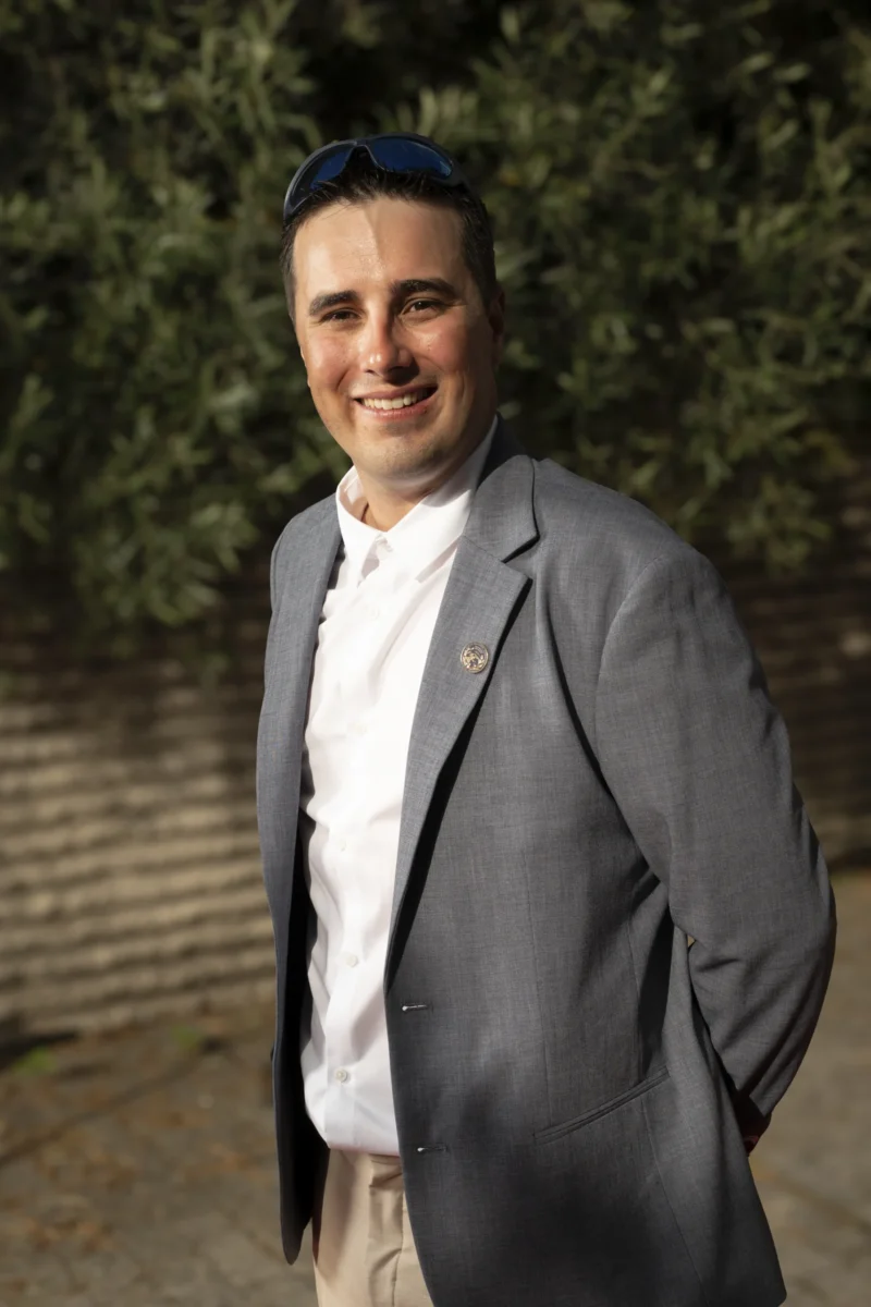 Cette photographie met en scène un homme souriant, sujet principal, vêtu d’un blazer gris, chemise blanche et kippa. Le cadrage est serré, centré sur lui, avec un arrière-plan flou de végétation.La lumière naturelle, douce et légèrement latérale, éclaire son visage, créant des ombres subtiles. Les couleurs dominantes sont les tons gris du blazer, le blanc de la chemise, et les verts naturels de l’arrière-plan.Le style est sobre et élégant, évoquant une ambiance calme et professionnelle, tout en dégageant une touche chaleureuse grâce au sourire du sujet.
