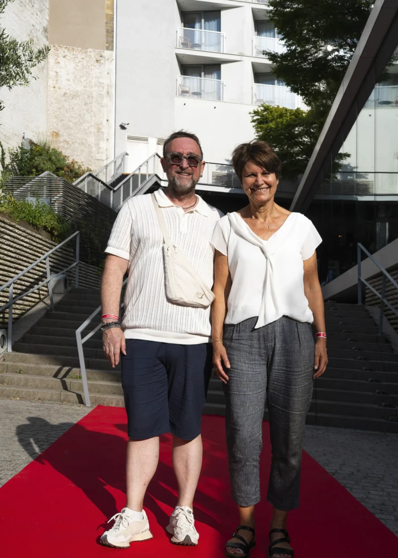 Cette photographie montre deux personnes posant sur un tapis rouge. Le sujet principal est ce couple souriant, l’homme en lunettes, t-shirt blanc et short, la femme en haut blanc et pantalon gris.La composition est symétrique, avec un cadrage serré sur eux. L’arrière-plan inclut des escaliers et un bâtiment moderne. La lumière est naturelle et douce, probablement en journée. Les couleurs dominantes sont les tons neutres (gris, blanc) et le rouge vif du tapis.Le style est simple et spontané, évoquant une ambiance détendue et élégante, typique d’un événement public ou culturel.