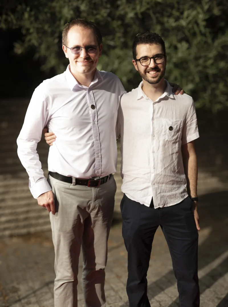 Cette photographie montre deux hommes posant côte à côte. Le sujet principal est leur présence amicale, avec un cadrage serré sur leurs bustes et épaules.La composition est symétrique et équilibrée. La lumière naturelle, douce et légèrement tamisée, semble provenir de la gauche, créant des ombres subtiles. Les couleurs dominantes sont des tons neutres : blanc (chemises), beige (pantalon), et verts discrets en arrière-plan.Le style est simple et naturel, évoquant une ambiance détendue et chaleureuse, presque intimiste.