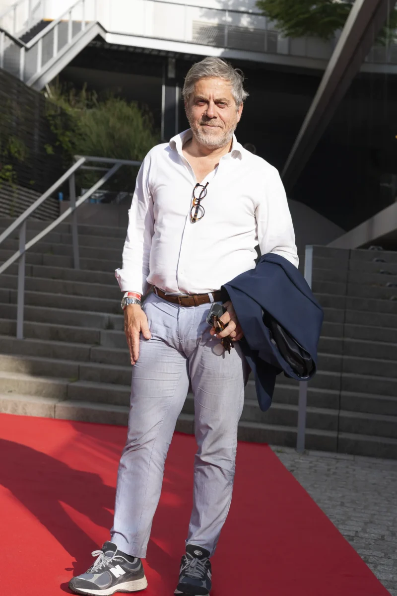 Cette photographie montre un homme comme sujet principal, souriant et détendu, vêtu d’une chemise blanche et d’un pantalon clair. Il tient une pochette à la main.La composition est simple et naturelle, avec un cadrage serré sur le sujet, mettant en valeur son expression. L’arrière-plan, des marches et une végétation, est flou, créant un effet de profondeur.La lumière semble naturelle et douce, éclairant bien le visage. Les couleurs dominantes sont les tons neutres (gris, blanc) et le rouge vif du tapis sous ses pieds.Le style est sobre et spontané, évoquant une ambiance décontractée et chaleureuse.