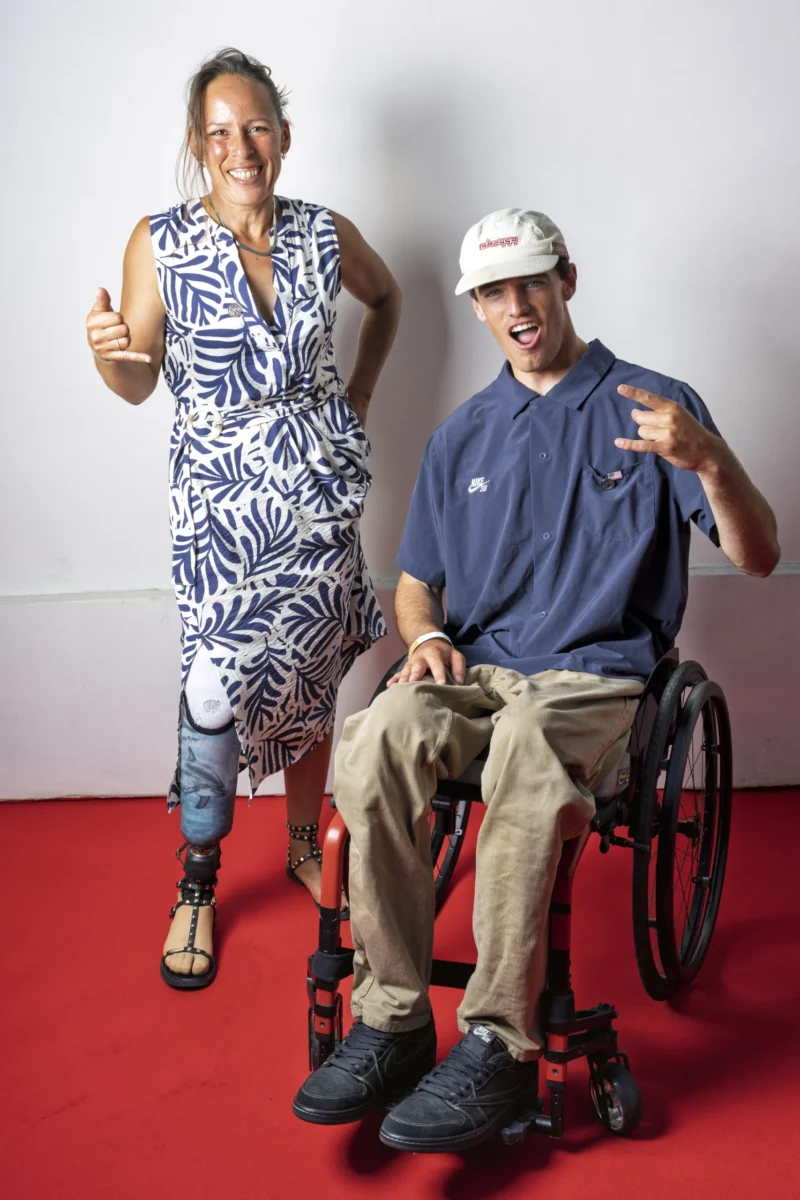 Cette photographie montre deux personnes souriantes, l’une debout et l’autre assise en fauteuil roulant. La femme porte une robe à motifs tropicaux, tandis que l’homme arbore un polo bleu et un chapeau.Le cadrage est serré, mettant en valeur leurs visages et leurs gestes positifs. La lumière est douce et uniforme, éclairant clairement les sujets. Les couleurs dominantes sont vives : bleu, rouge (sol), beige et motifs colorés.L’ambiance est joyeuse et inclusive, reflétant une énergie positive et une connexion chaleureuse.