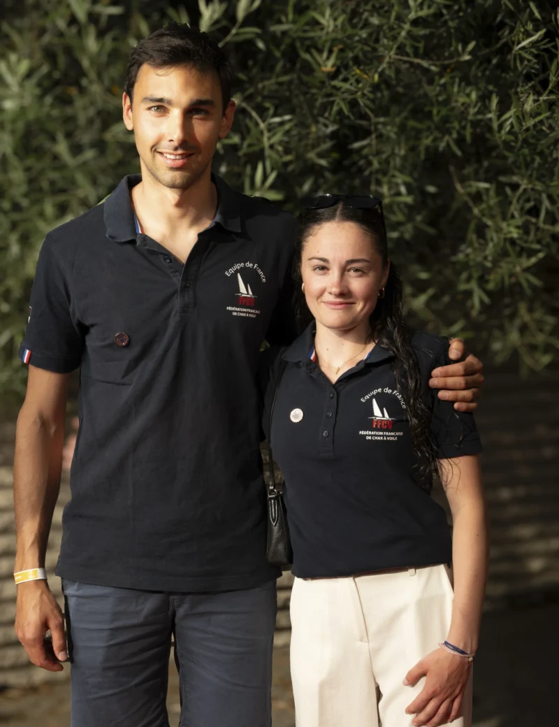 Cette photographie montre deux jeunes posant côte à côte. Le sujet principal est un couple souriant, vêtu de polo identiques avec un logo.La composition est symétrique et serrée, avec un cadrage en plan moyen. L'arrière-plan flou met en valeur les sujets grâce à un effet bokeh. La lumière naturelle, douce et légèrement latérale, éclaire leurs visages, créant des ombres subtiles. Les couleurs dominantes sont les tons bleus des polos, blancs des vêtements et verts des feuilles en arrière-plan.Le style est simple et naturel, évoquant une ambiance chaleureuse et conviviale.
