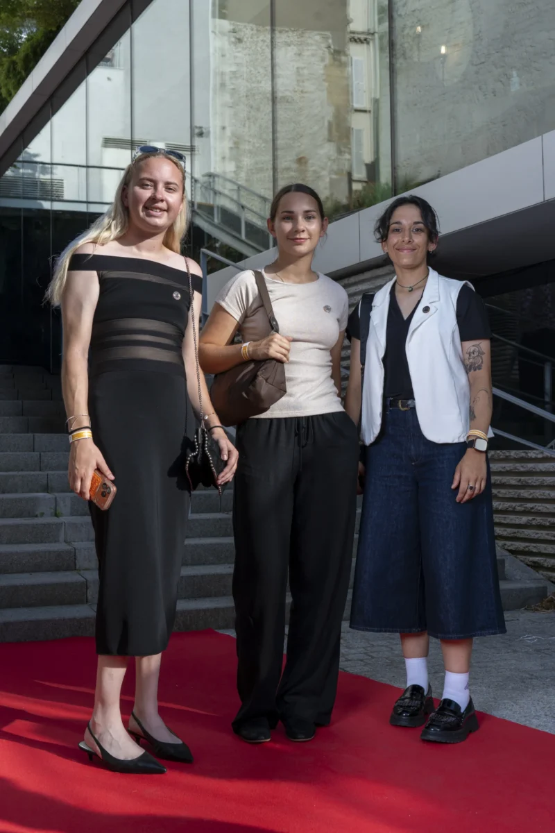 Cette photographie montre trois femmes posant sur un tapis rouge. Le sujet principal est leur présence ensemble, avec une composition centrée et symétrique. Le cadrage est serré, mettant en valeur leurs tenues élégantes.La lumière est naturelle et douce, éclairant leurs visages. Les couleurs dominantes sont les tons neutres (noir, blanc, bleu) avec des touches de rouge sur le tapis. Le style est sophistiqué et urbain, évoquant un événement formel. L’ambiance est chic et conviviale.