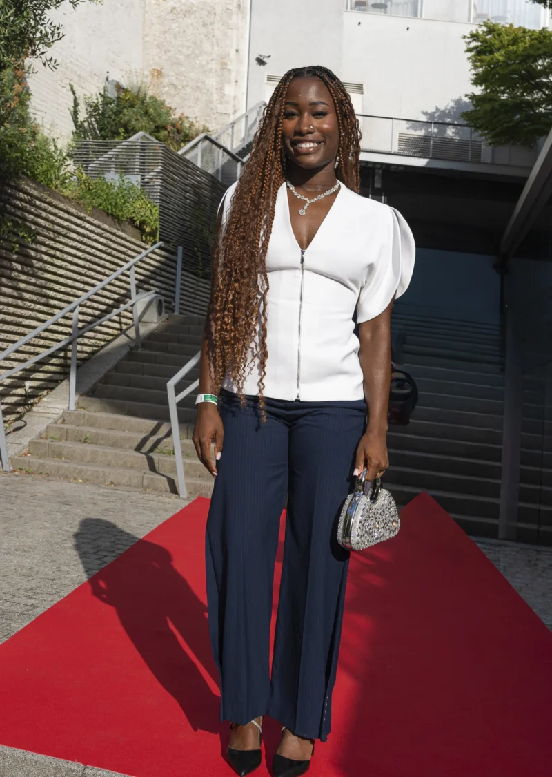 Cette photographie met en scène une femme comme sujet principal, debout sur un tapis rouge. Elle porte une tenue élégante : haut blanc et pantalon bleu foncé, avec des accessoires discrets.La composition est simple et centrée, avec un cadrage serré sur elle. L’arrière-plan montre des marches en pierre et une structure architecturale sobre. La lumière est naturelle et douce, éclairant son visage sans ombres marquées. Les couleurs dominantes sont le blanc, le bleu et le rouge du tapis, créant une ambiance chic et professionnelle. Le style est élégant et contemporain, évoquant un événement formel.