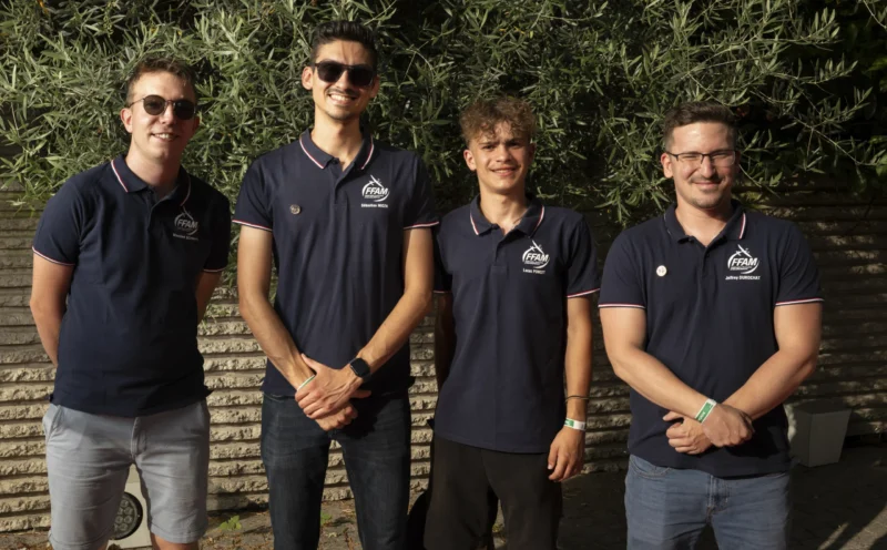Cette photographie montre quatre jeunes hommes posant côte à côte. Le sujet principal est leur groupe, habillés de polos bleus identiques, suggérant une appartenance commune (équipe ou club).La composition est symétrique, avec un cadrage serré sur les épaules et le haut du corps. La lumière naturelle, douce et homogène, éclaire le groupe sans ombres marquées. Les couleurs dominantes sont les bleus des polos et les verts des feuillages en arrière-plan.Le style est sobre et professionnel, évoquant une ambiance conviviale et unifiée, renforcée par leur posture détendue et souriante.
