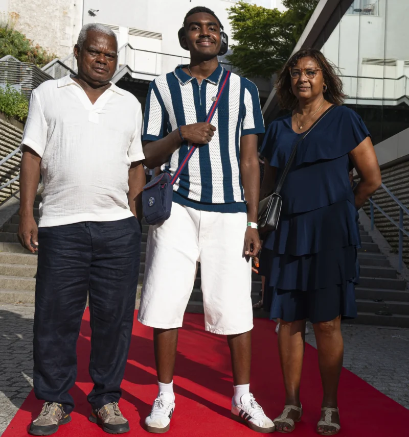 Cette photographie met en scène trois personnes posant sur un tapis rouge. Le sujet principal semble être le jeune homme au centre, vêtu d’un polo rayé et d’un short blanc, encadré par deux adultes souriants.La composition est symétrique, avec un cadrage serré sur les trois individus. La lumière est naturelle et douce, éclairant uniformément les visages. Les couleurs dominantes sont les tons neutres (blanc, bleu, gris) et les touches de bleu foncé et de rouge (tapis).Le style est sobre et formel, évoquant une ambiance de cérémonie ou d’événement public. L’ambiance est chaleureuse et familiale.