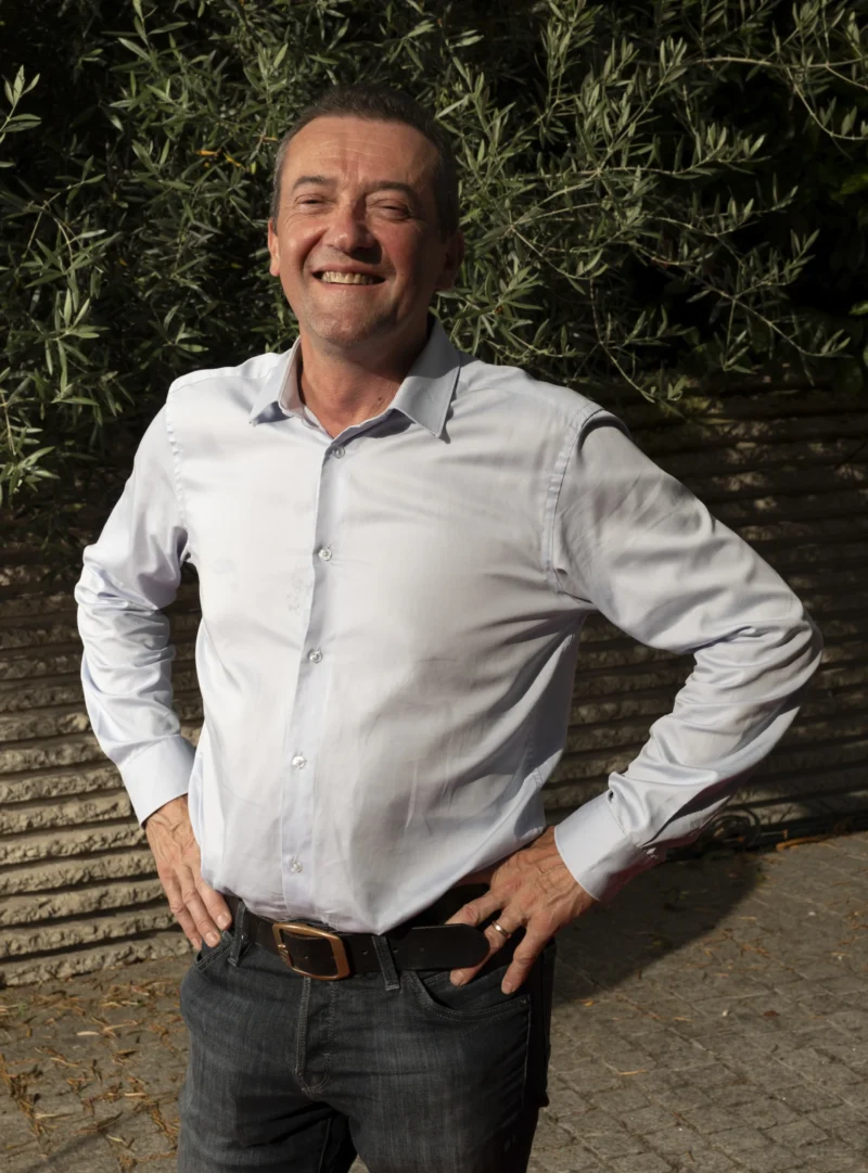 Cette photographie met en scène un homme souriant comme sujet principal, vêtu d'une chemise blanche et d'un jean. Le cadrage est serré, centré sur lui, avec un fond légèrement flou de végétation.La lumière naturelle éclaire son visage, créant des ombres douces. Les couleurs dominantes sont les tons neutres (blanc, gris du jean) et les verts de la végétation en arrière-plan. Le style est simple et naturel, évoquant une ambiance détendue et chaleureuse, presque informelle.