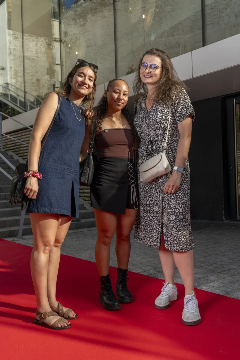 Cette photographie montre trois femmes posant ensemble sur un tapis rouge. Le sujet principal est leur complicité et leur élégance.La composition est symétrique, avec les trois femmes alignées. Le cadrage est serré, mettant en valeur leurs visages et tenues. La lumière est douce et naturelle, éclairant bien leurs traits.Les couleurs dominantes sont des tons neutres (noir, gris) et des touches de couleurs vives (robe noire, imprimé floral, accessoires colorés). Le style est chic et moderne, évoquant une ambiance festive et sophistiquée, probablement lors d’un événement mondain.