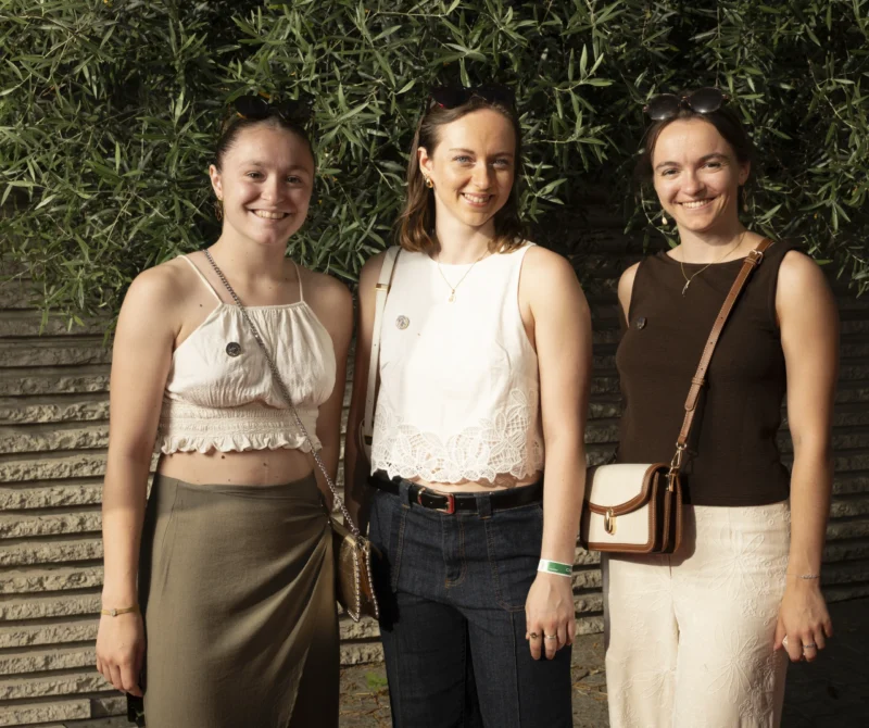 Cette photographie montre trois femmes souriantes posant côte à côte. Le sujet principal est leur complicité et leur harmonie.La composition est symétrique, avec un cadrage serré sur elles. L’arrière-plan flou, composé de feuillage, isole les sujets. La lumière naturelle, douce et légèrement tamisée, crée une ambiance chaleureuse.Les couleurs dominantes sont des tons neutres : beige, blanc et vert. Le style est décontracté et élégant, évoquant une ambiance conviviale et détendue, probablement en extérieur lors d’un moment informel.
