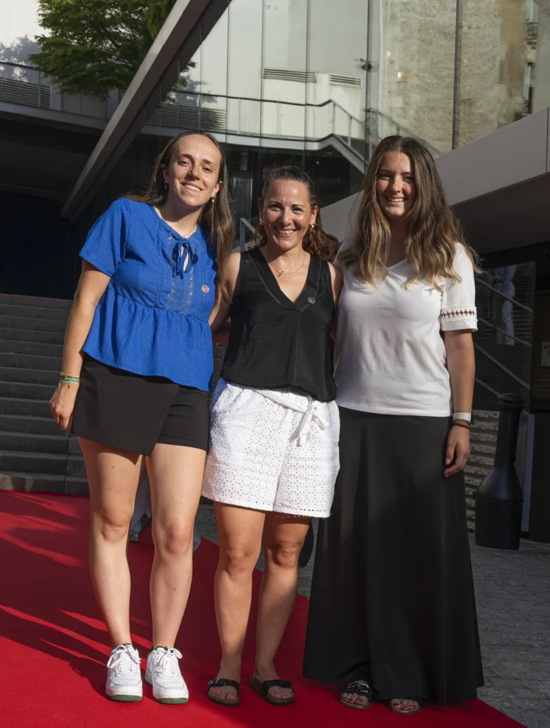 Cette photographie montre trois femmes souriantes posant sur un tapis rouge. Le sujet principal est leur complicité et leur joie apparente.La composition est symétrique, avec les trois femmes alignées côte à côte. Le cadrage est serré, mettant en valeur leurs visages et le haut de leur corps. La lumière est naturelle et douce, éclairant uniformément les sujets.Les couleurs dominantes sont les tons clairs : blanc, bleu et noir, avec une touche de rouge due au tapis. Le style est décontracté et élégant, évoquant une ambiance conviviale et festive, probablement lors d’un événement public.