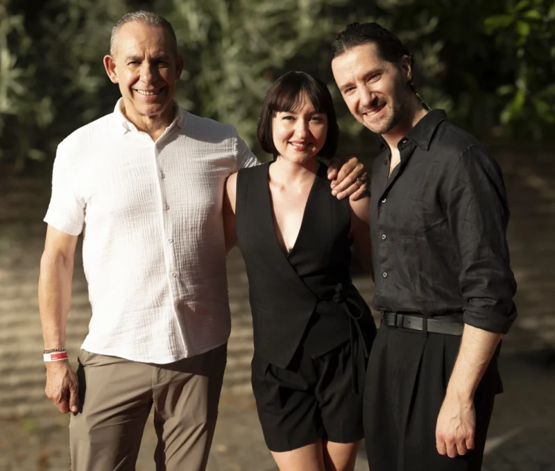Cette photographie montre trois personnes posant ensemble. Le sujet principal est ce trio, composé d’un homme âgé à gauche, et de deux femmes plus jeunes au centre et à droite.La composition est symétrique et harmonieuse, avec un cadrage serré sur les visages. La lumière naturelle, douce et légèrement latérale, adoucit les traits et crée des ombres subtiles. Les couleurs dominantes sont neutres : tons beiges, gris et noirs, avec des touches de vert en arrière-plan.Le style est sobre et naturel, évoquant une ambiance chaleureuse et intime, presque familiale.