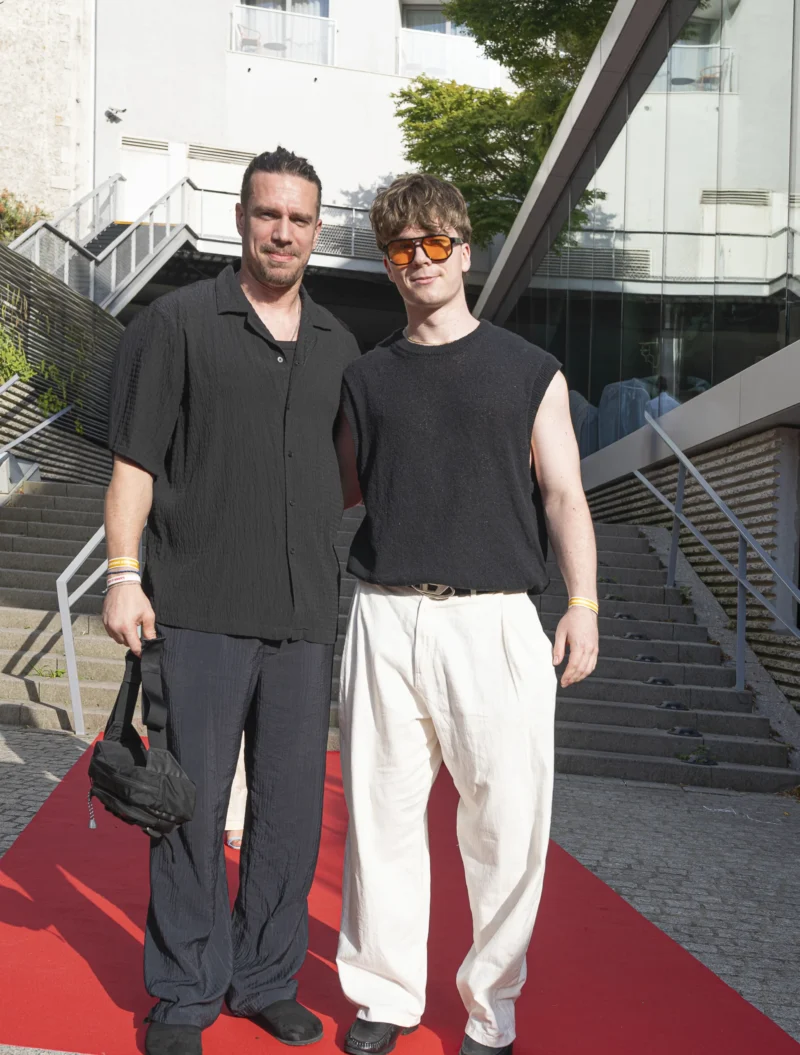 Cette photographie montre deux hommes posant côte à côte sur un tapis rouge. Le sujet principal est leur présence, habillés de manière décontractée mais élégante.La composition est symétrique, avec un cadrage serré sur les deux individus. L’arrière-plan architectural moderne et les escaliers métalliques ajoutent un contraste urbain. La lumière semble naturelle, légèrement douce, sans ombres marquées. Les couleurs dominantes sont les tons neutres (noir, gris, blanc) avec une touche de rouge du tapis.L’ambiance est à la fois chic et décontractée, évoquant un événement public ou une sortie informelle en milieu urbain.