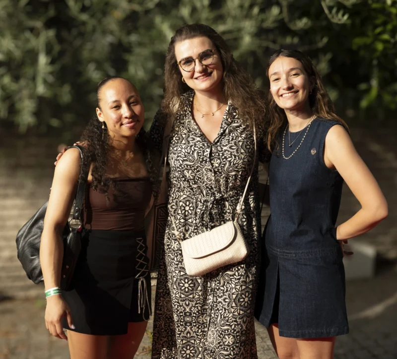 Cette photographie montre trois femmes souriantes posant côte à côte. Le sujet principal est leur complicité et leur harmonie.La composition est symétrique, avec un cadrage serré sur elles. La lumière est douce et naturelle, probablement en extérieur, créant des ombres légères. Les couleurs dominantes sont des tons neutres et naturels : noir, gris, bleu marine et touches de vert en arrière-plan.Le style est décontracté et élégant, évoquant une ambiance chaleureuse et amicale.