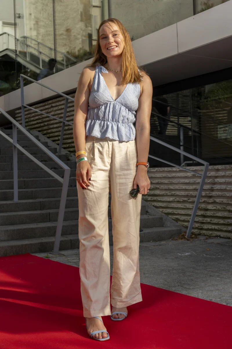 Cette photographie met en scène une jeune femme comme sujet principal, debout sur un tapis rouge. Elle porte une tenue élégante composée d’un haut bleu clair et de pantalons larges beige.La composition est simple et centrée, avec un cadrage serré sur elle, mettant en valeur sa silhouette. L’arrière-plan, des escaliers et une structure architecturale moderne, suggère un événement formel. La lumière est naturelle et douce, éclairant son visage sans ombres marquées.Les couleurs dominantes sont les tons pastel (bleu clair, beige) et le rouge du tapis, créant une ambiance chic et sophistiquée. Le style est à la fois décontracté et élégant.