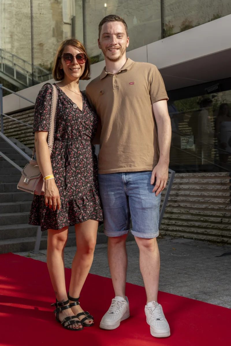Cette photographie montre un couple souriant sur un tapis rouge. Les sujets principaux sont une femme vêtue d’une robe fleurie et d’un homme en polo beige et short, posant côte à côte.La composition est symétrique, avec un cadrage serré sur eux. L’arrière-plan, légèrement flou, révèle des escaliers et une structure architecturale moderne. La lumière est naturelle et douce, éclairant uniformément les visages. Les couleurs dominantes sont les tons pastel (beige, rose pâle) et les motifs floraux.Le style est décontracté et élégant, évoquant une ambiance festive ou un événement public.