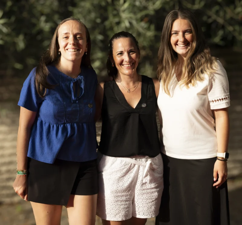 Cette photographie met en scène trois femmes souriantes posant côte à côte. Le sujet principal est leur complicité et leur harmonie. La composition est symétrique, avec un cadrage serré sur elles, en pied et légèrement en contre-plongée. La lumière naturelle, douce et diffuse, provient probablement du jour, créant des ombres légères. Les couleurs dominantes sont des tons pastel : bleu, noir et blanc, avec des touches de vert en arrière-plan. Le style est simple et naturel, évoquant une ambiance chaleureuse et conviviale, presque familiale ou amicale.
